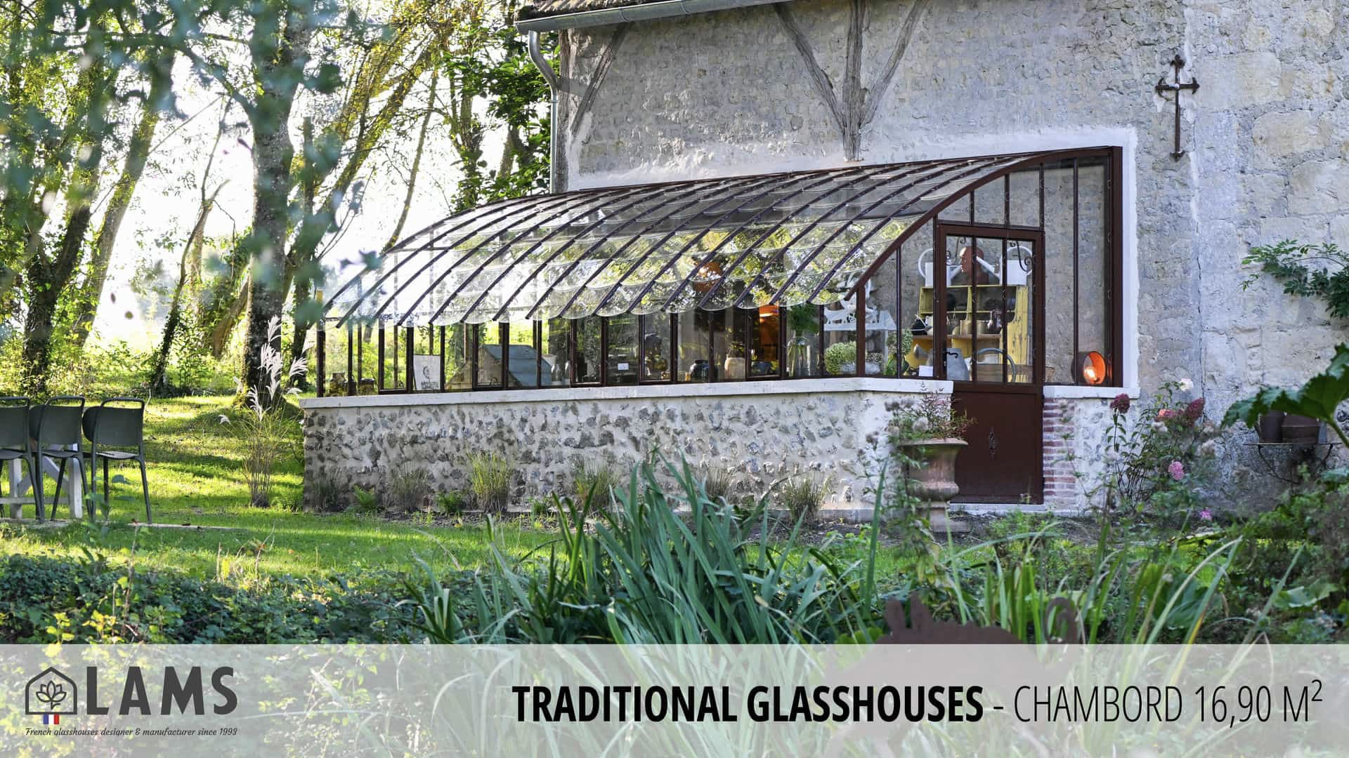 A traditional glasshouse with wooden frames sits in a lush garden. Stone building exterior and empty chairs nearby suggest tranquility.