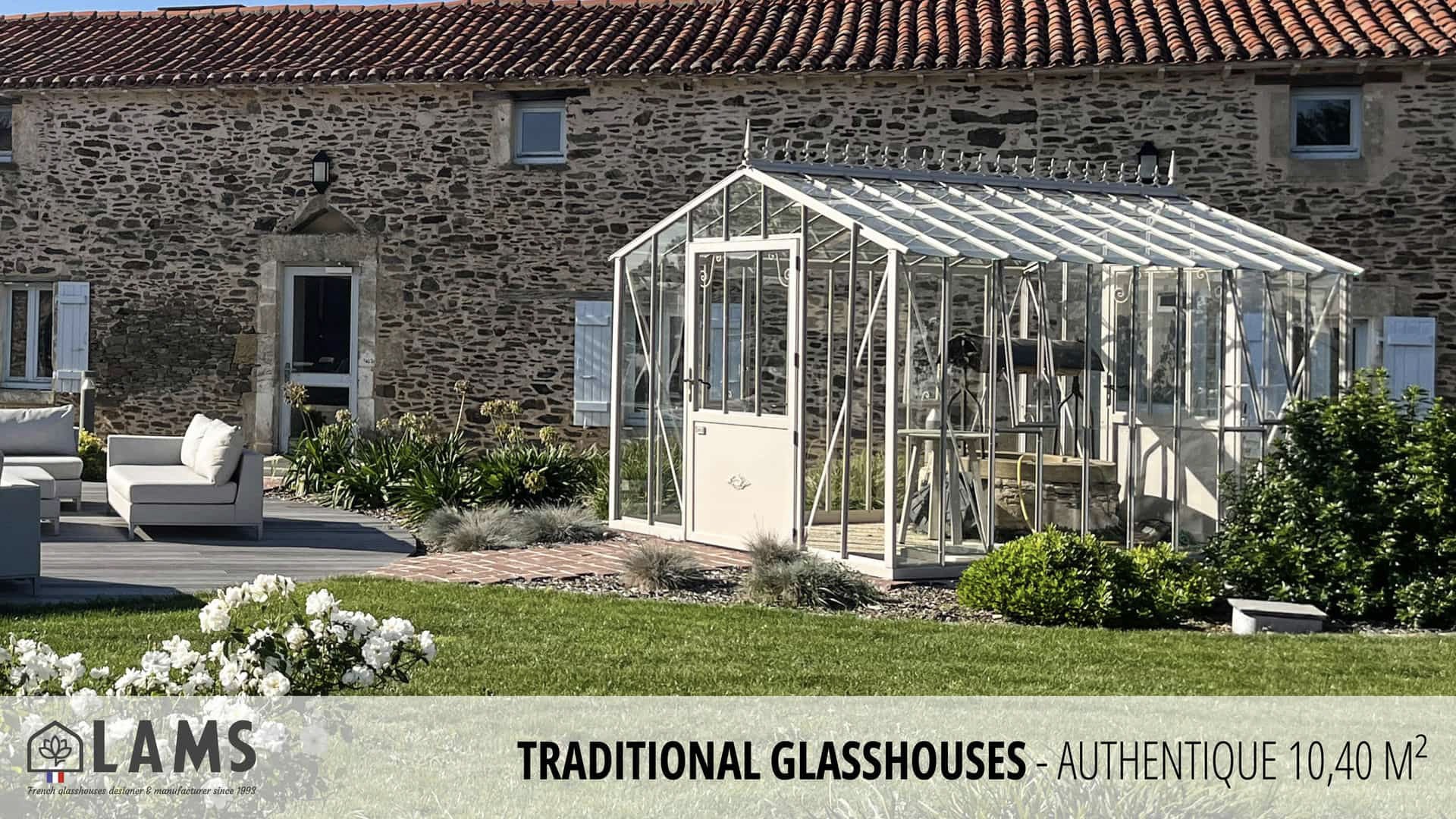 A traditional glasshouse is set in a landscaped garden with a stone building and outdoor seating area nearby.