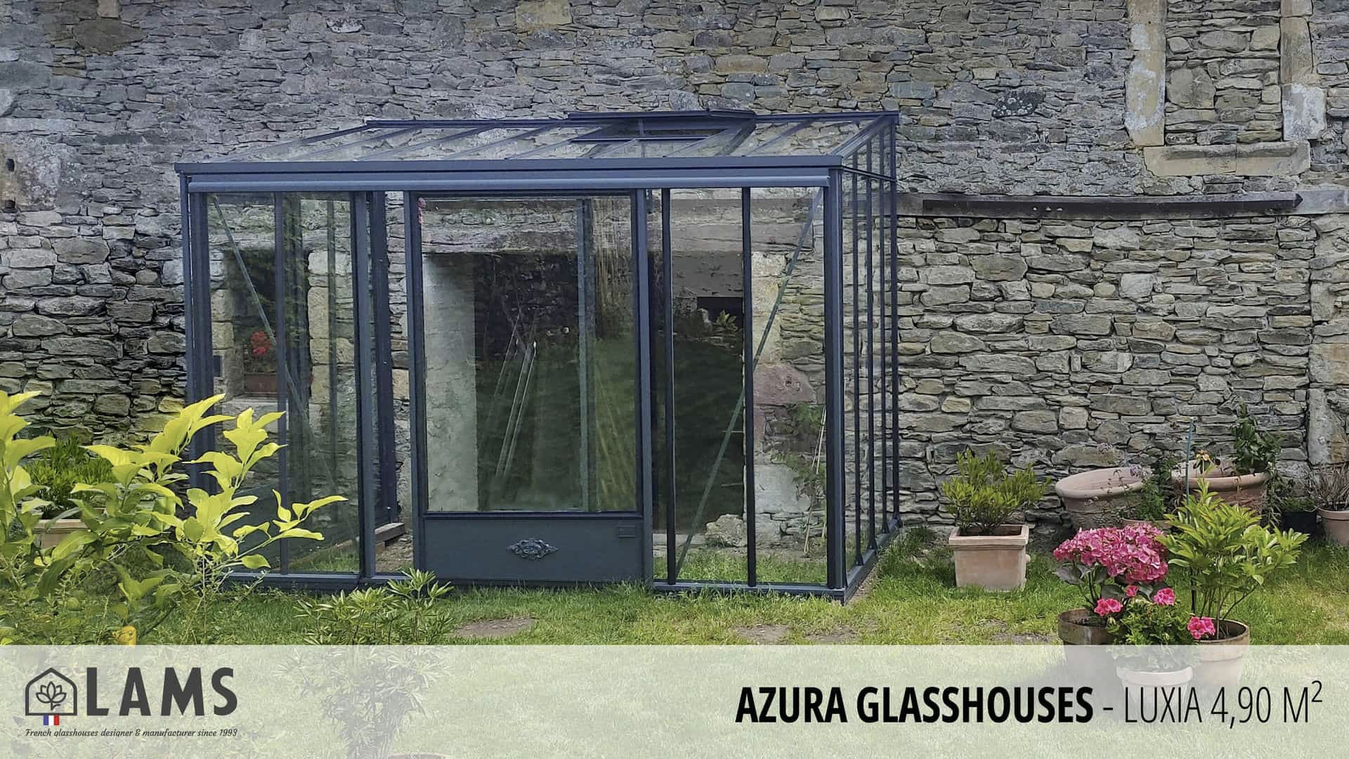 A small glasshouse stands in a lush garden against a stone wall, surrounded by potted plants and grass.