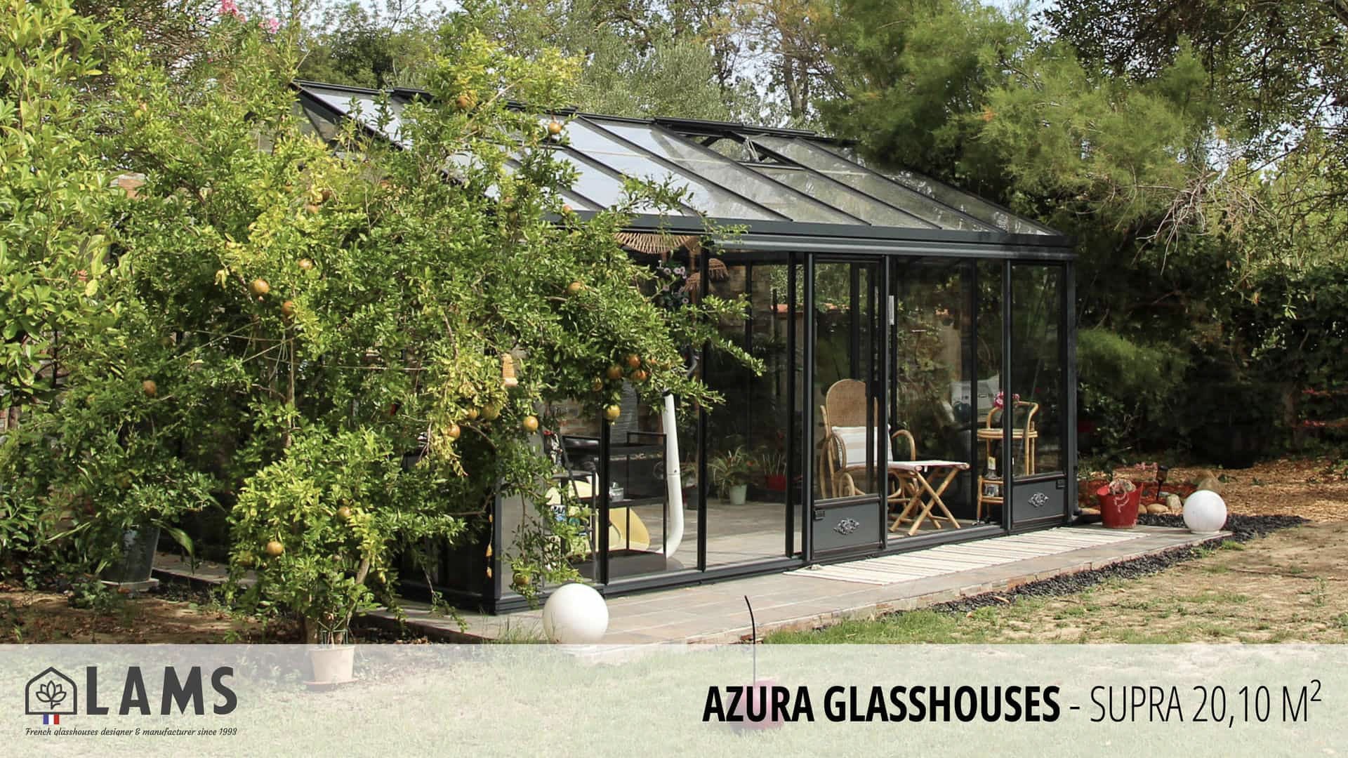 A modern glasshouse surrounded by lush greenery, featuring a chair and table inside. Sunlight filters through the transparent structure.