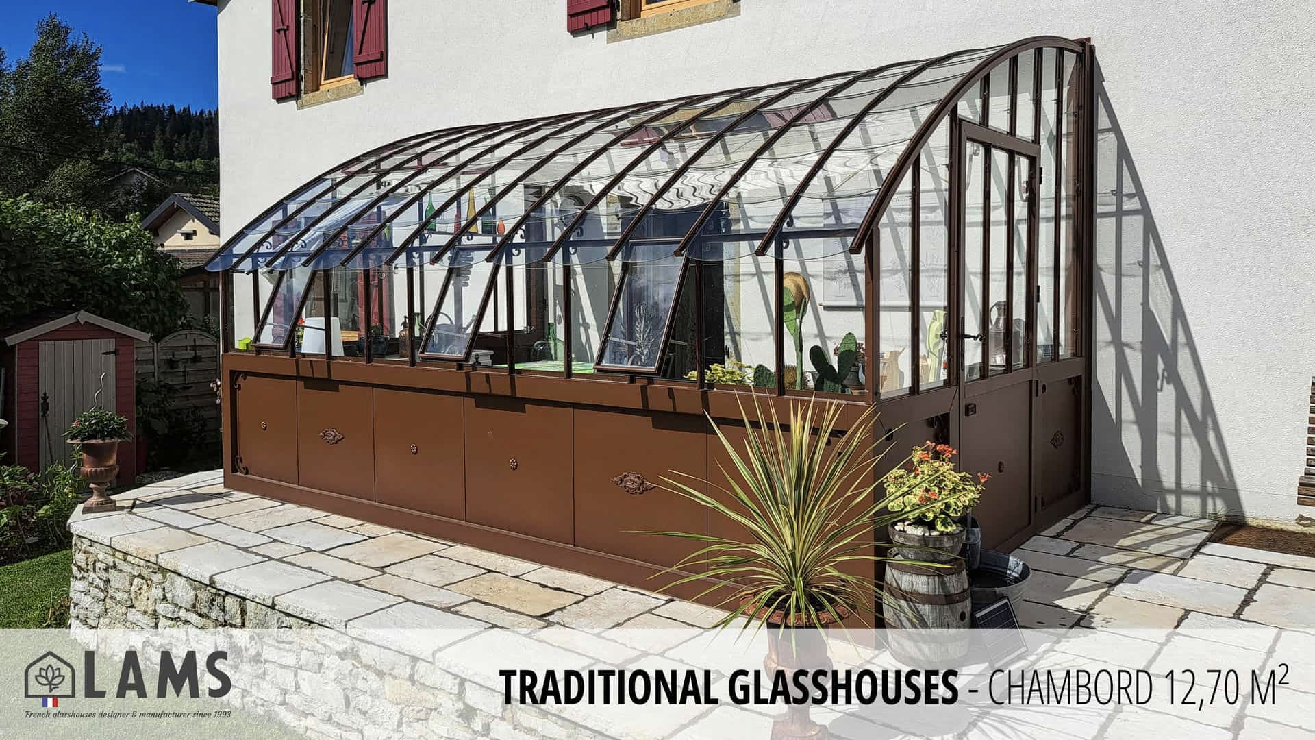 A traditional glasshouse attached to a building, surrounded by a stone patio. Plants and a small shed are nearby.