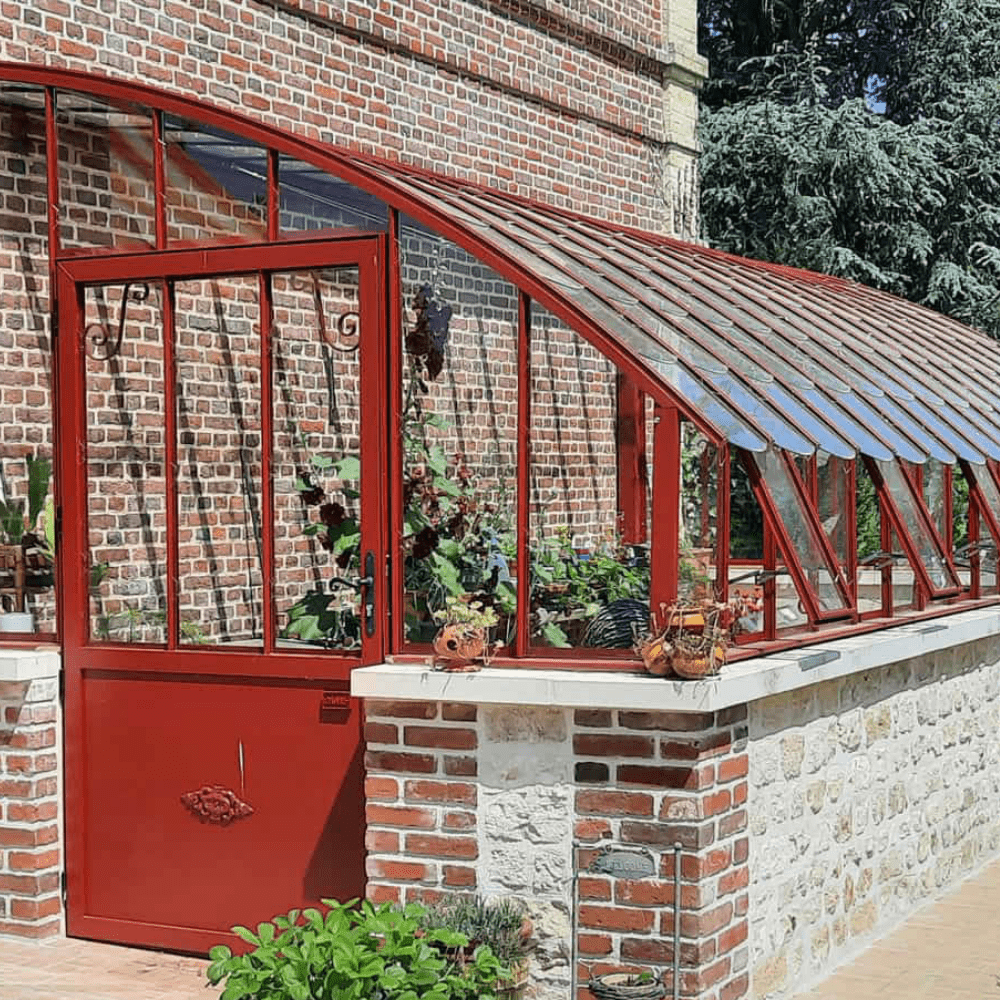 A red-framed greenhouse is attached to a brick building. It features glass panels, plants inside, and decorative items outside, surrounded by greenery.