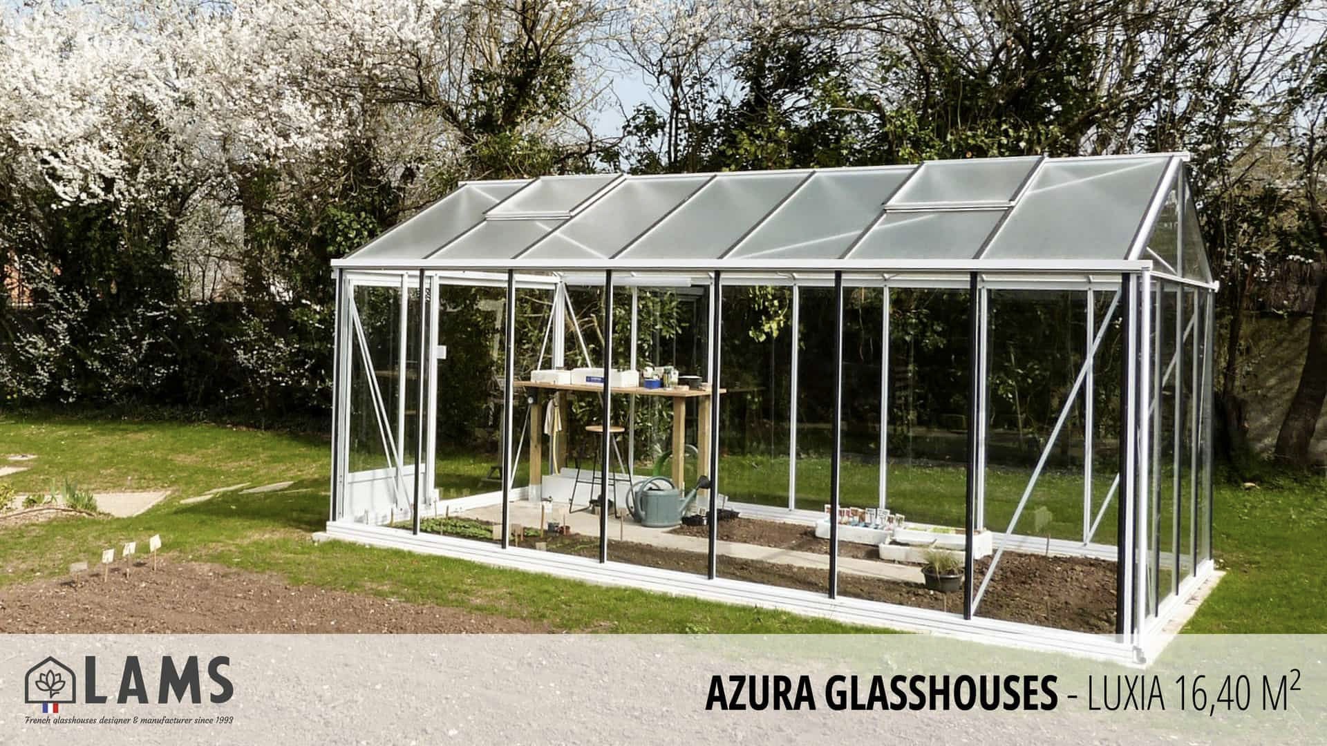 A glass greenhouse in a garden setting with blooming trees and a labeling for Azura Glasshouses - Luxia 16.40 m².