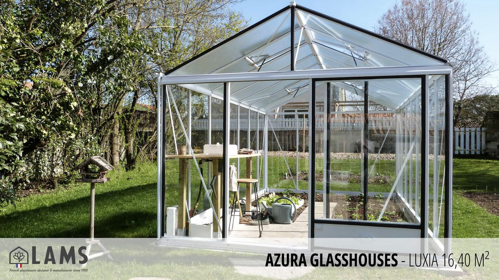 A modern glasshouse stands in a garden, surrounded by green grass and trees. Gardening tools and plants are visible inside.