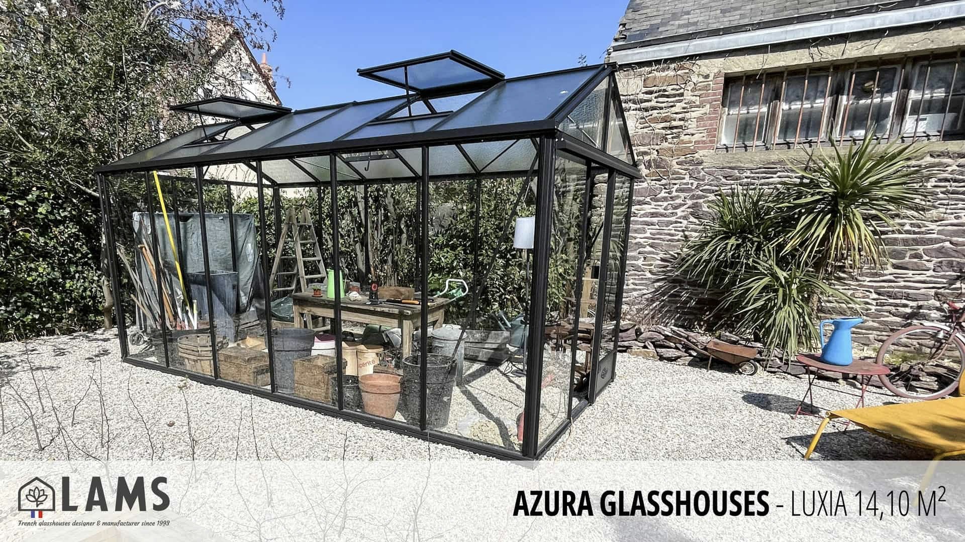 A glasshouse stands in a garden area beside a stone building, with various gardening tools and plants visible through its transparent walls.