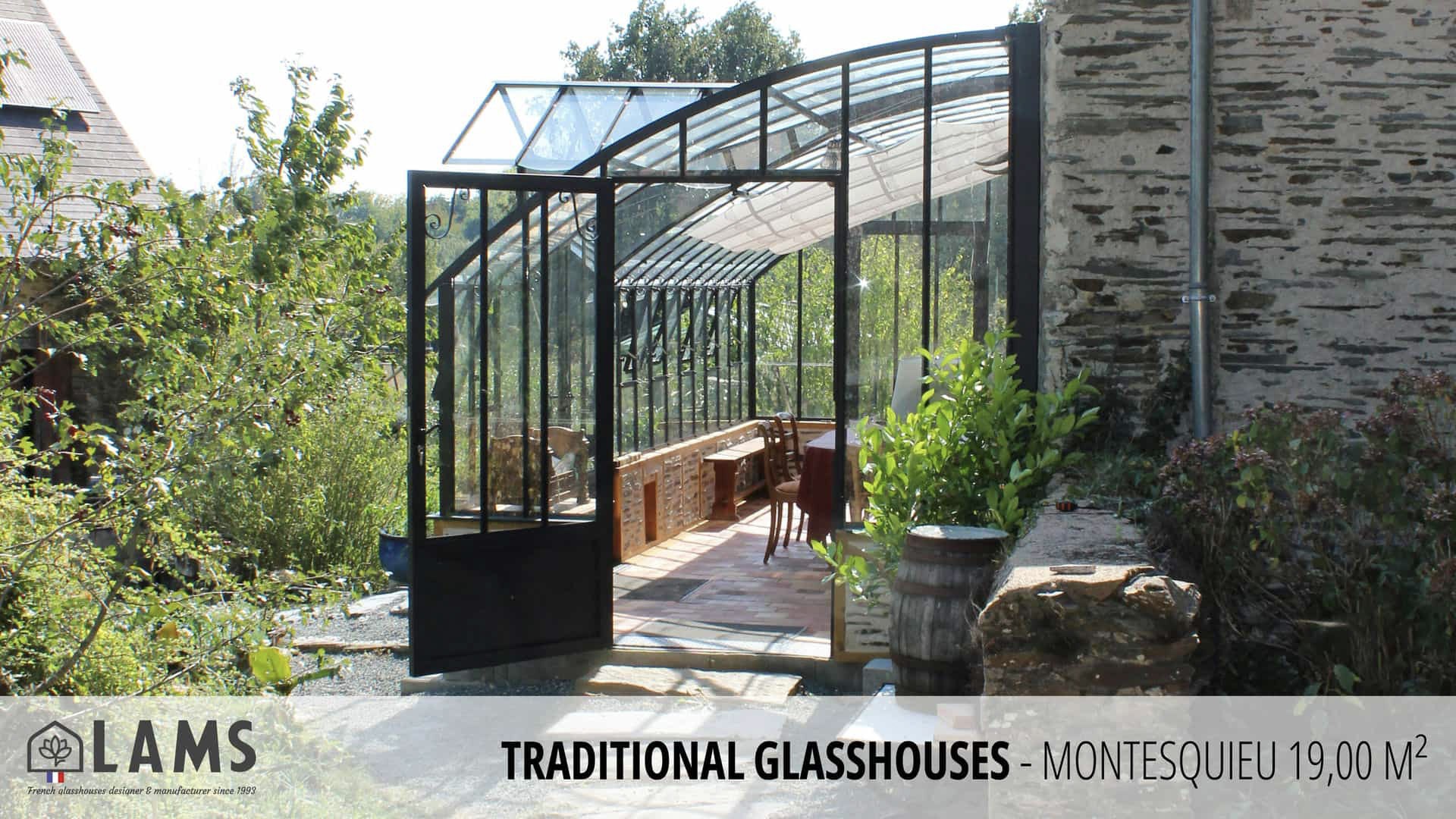 A traditional glasshouse with an open door, surrounded by lush greenery and a stone wall in a garden setting.
