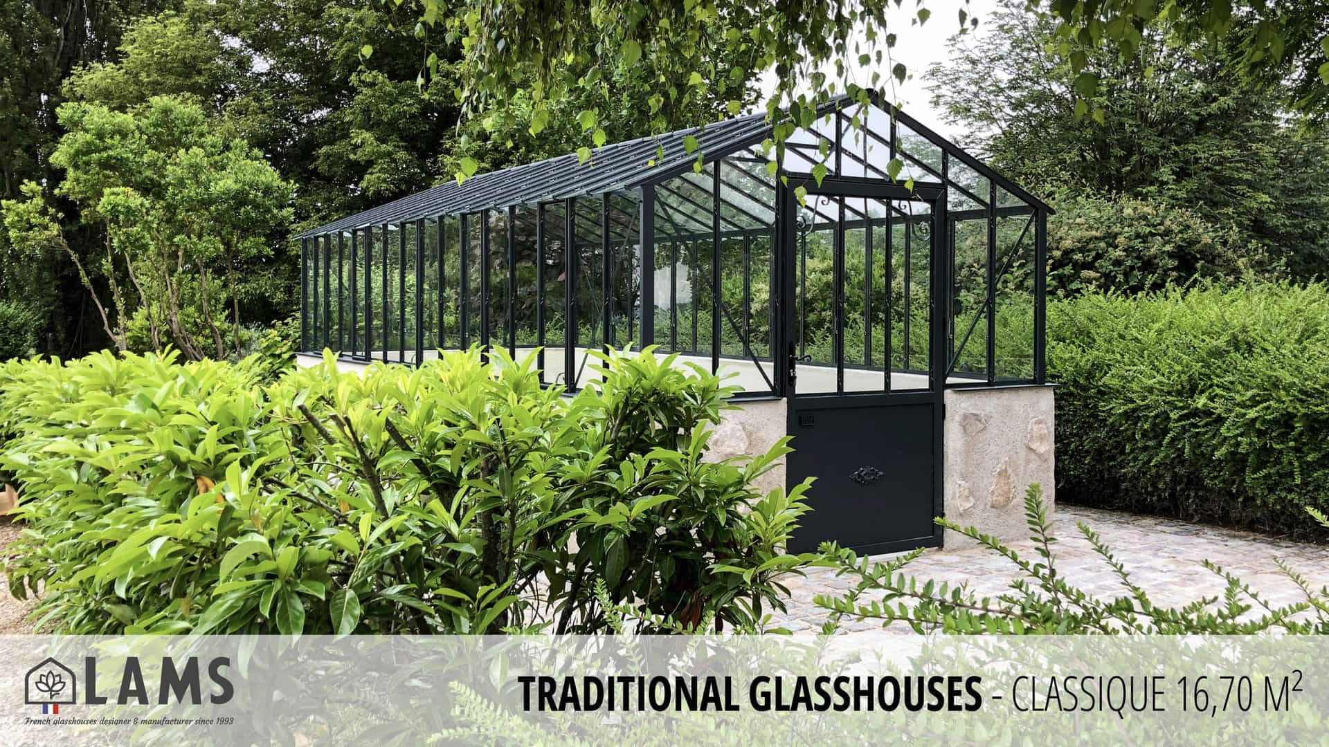 A traditional glasshouse with a black frame stands amidst lush greenery. The scene is peaceful and natural, with a clear pathway.