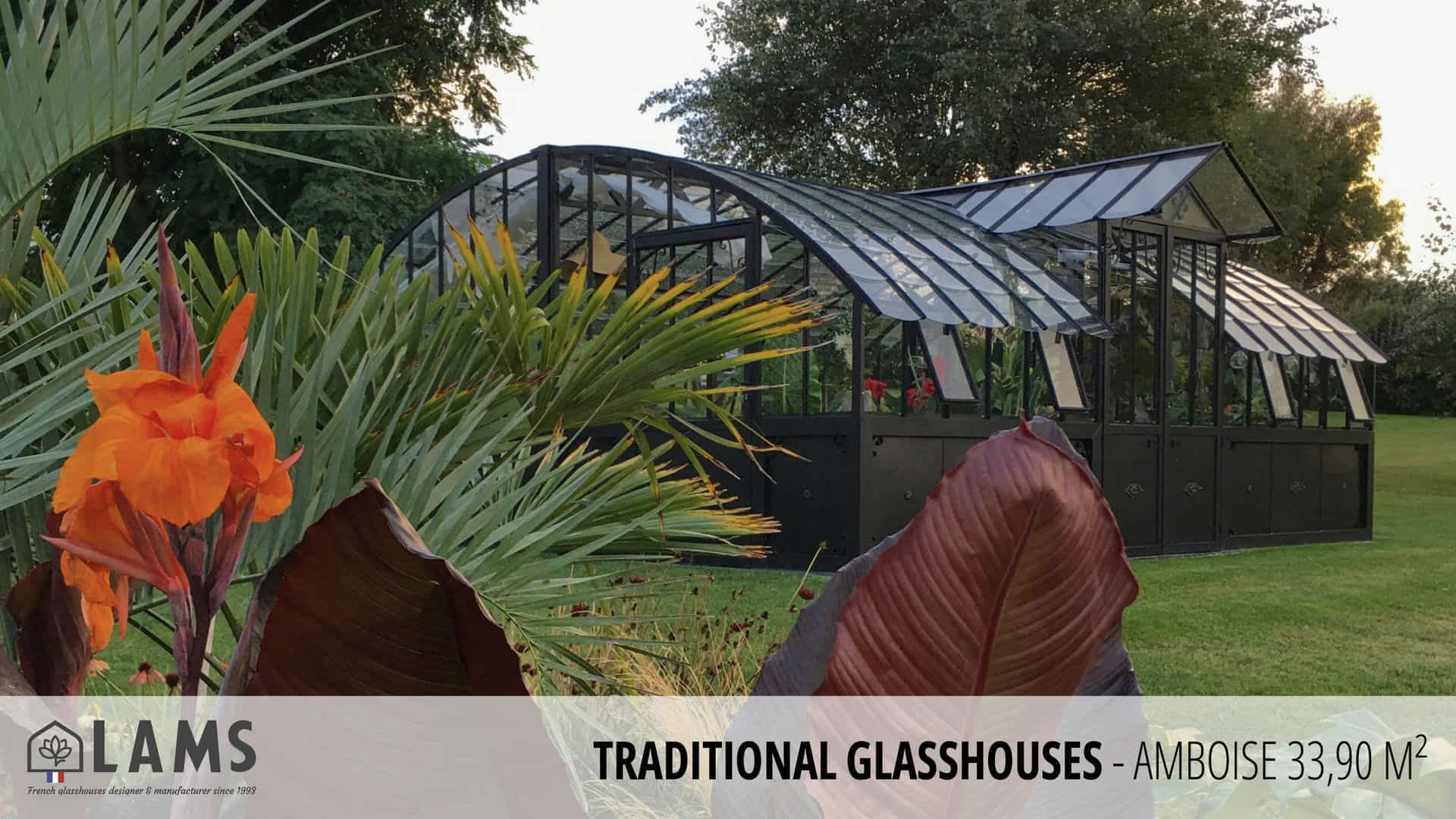 A traditional glasshouse with arched, metal framework is surrounded by lush green grass and colorful plants under a clear sky.