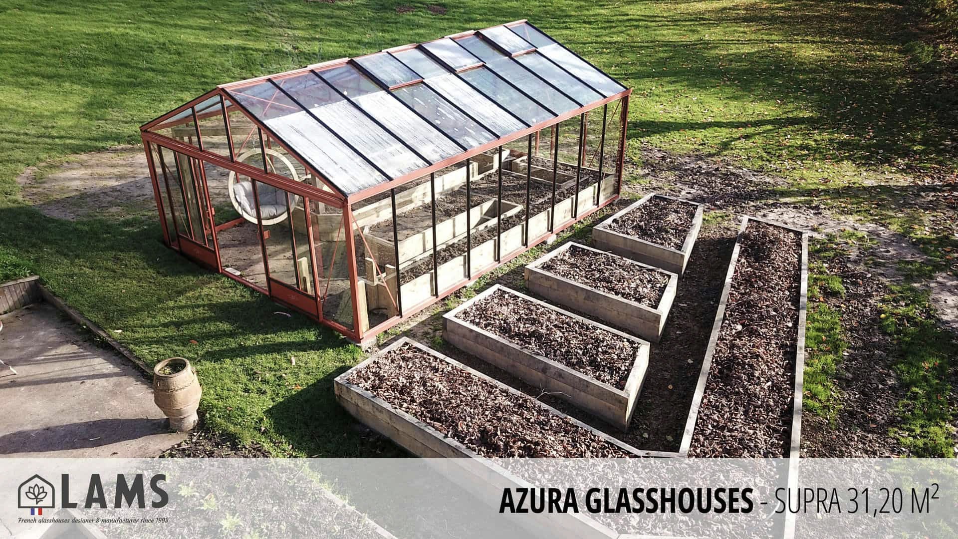 A glass greenhouse with adjacent raised garden beds, set in a grassy area. No people or recognizable landmarks are present.