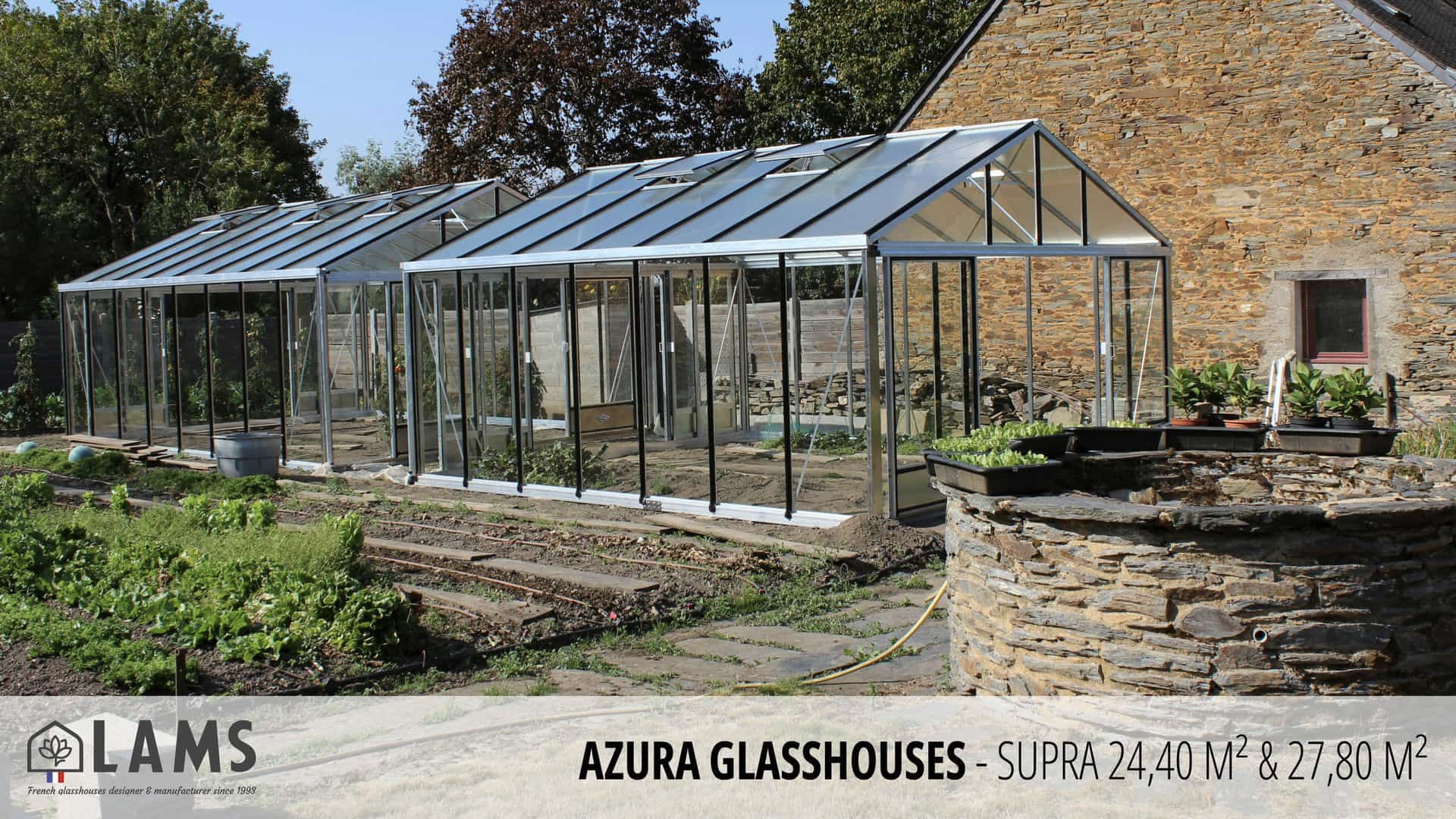 A spacious garden features Azura glasshouses beside a stone building, with lush greenery and stone-lined paths under clear skies.