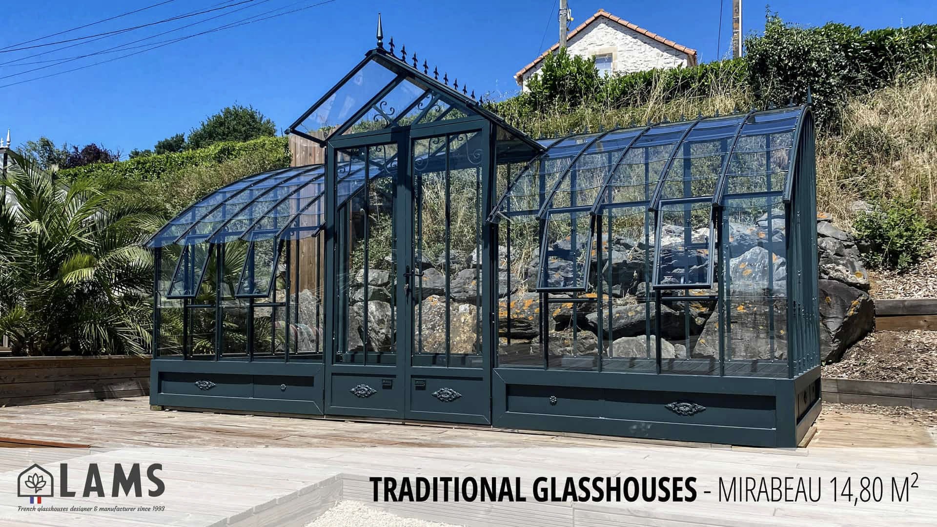A traditional glasshouse with a black frame sits in a sunny, landscaped area, surrounded by greenery and a stone wall backdrop.