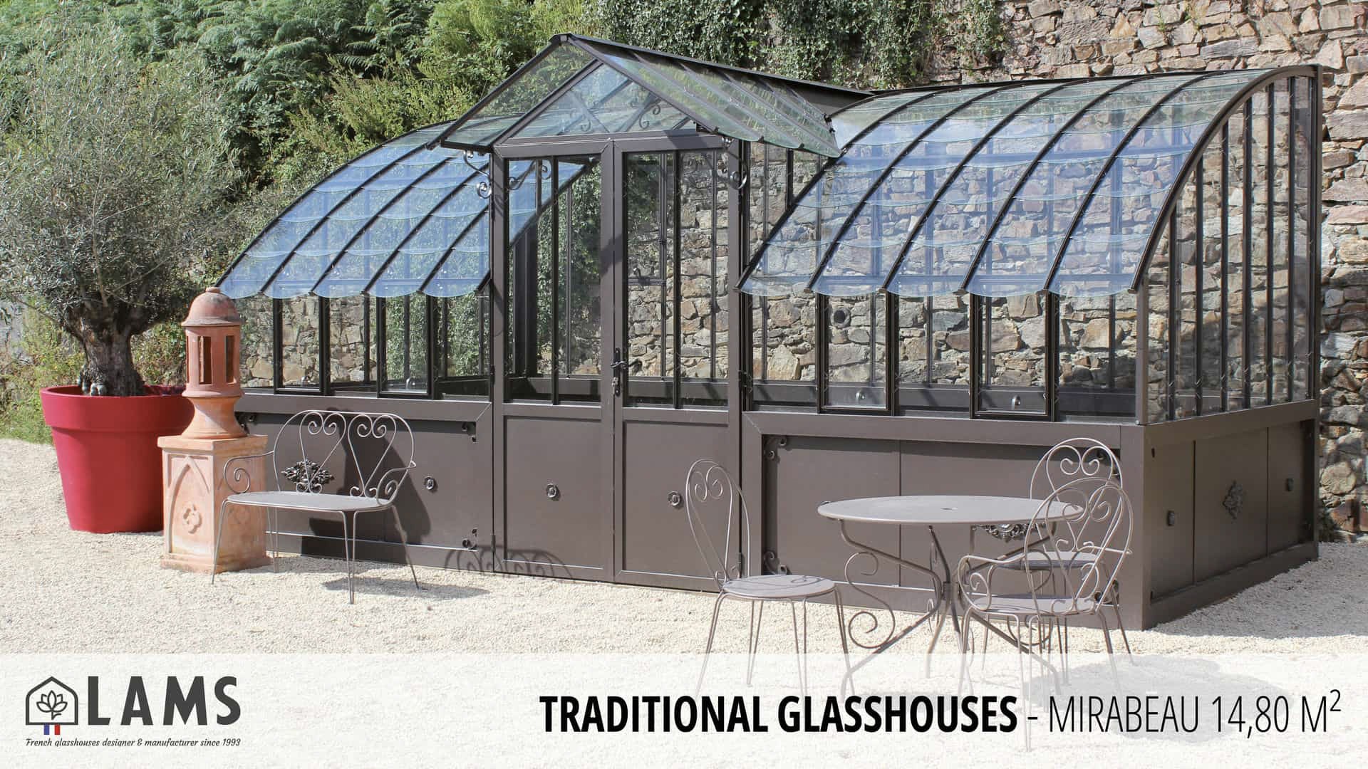A traditional glasshouse with curved roof sits beside wrought iron furniture, framed by a stone wall and lush greenery, under a clear sky.