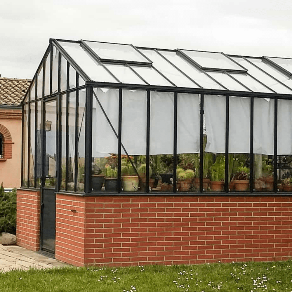A quaint greenhouse with brick base stands near a terracotta-colored house with blue shutters, surrounded by landscaped garden and potted plants.