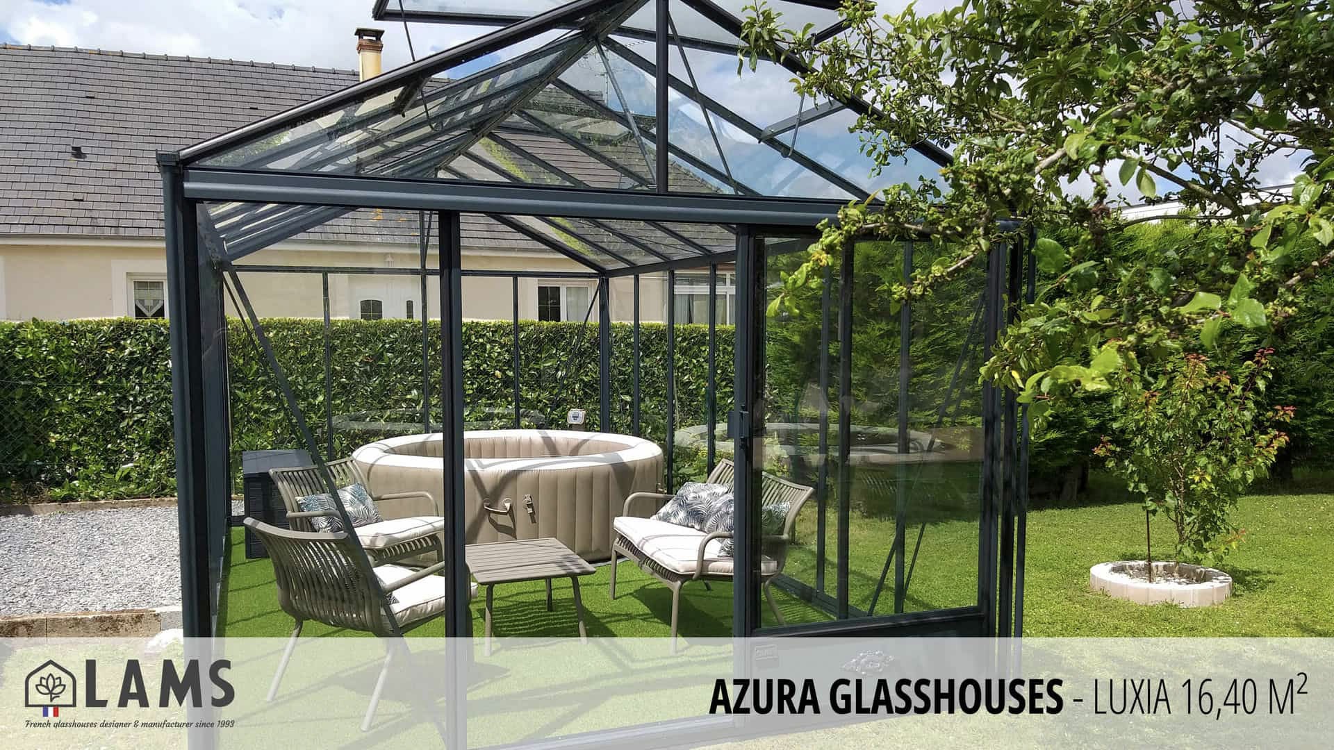 A modern glasshouse contains a cozy seating area with chairs and a cushioned round seat, surrounded by greenery and a lawn.