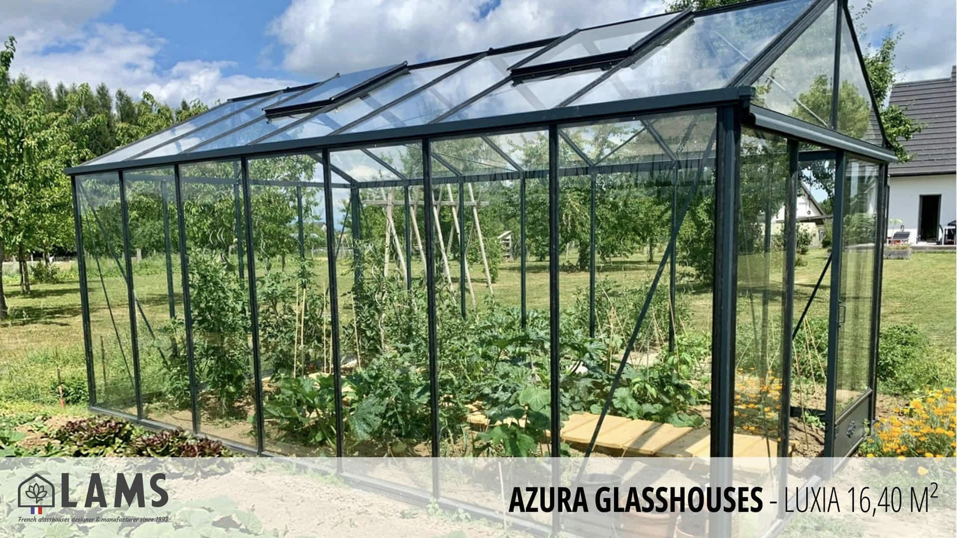 A glass greenhouse with plants inside, situated in a lush garden. A house and trees are visible in the background under a blue sky.