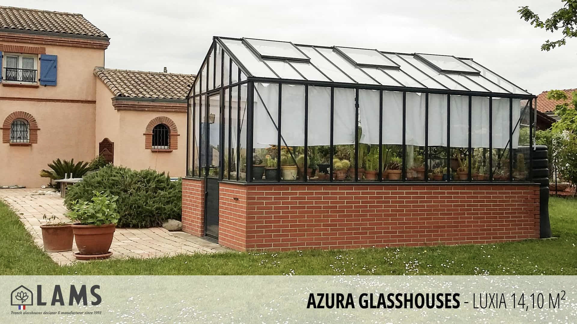 A glasshouse with lush plants stands beside a charming rustic building. The yard has potted plants and a paved pathway.