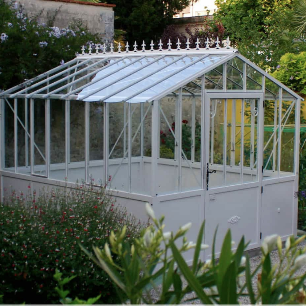 A small, white greenhouse with decorative trim stands in a lush garden surrounded by flowering plants and greenery, under a clear sky.